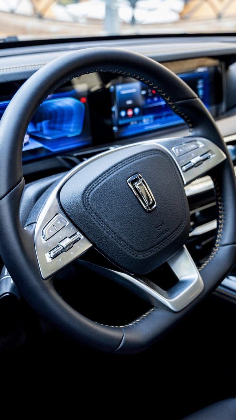 Volante moderno de cuero negro con botones plateados y una pantalla digital azul de fondo.