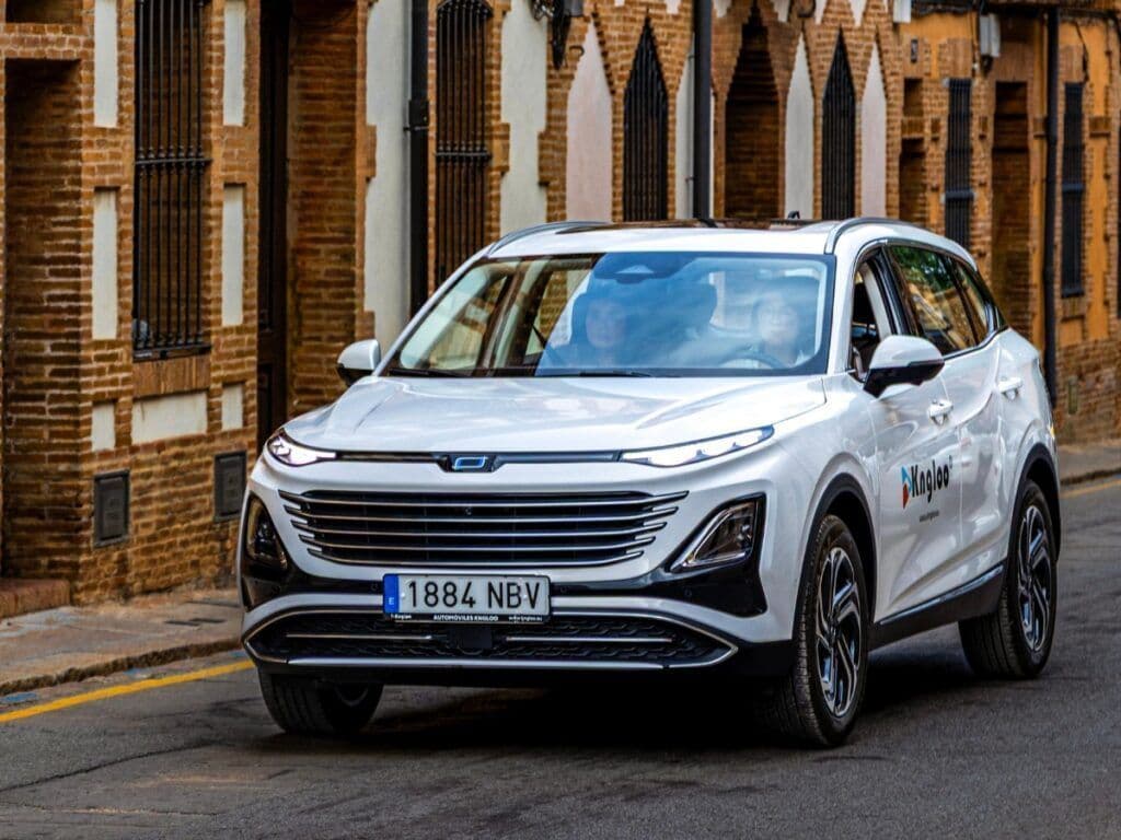 SUV blanco con luces encendidas circulando por una calle flanqueada por edificios de ladrillo.